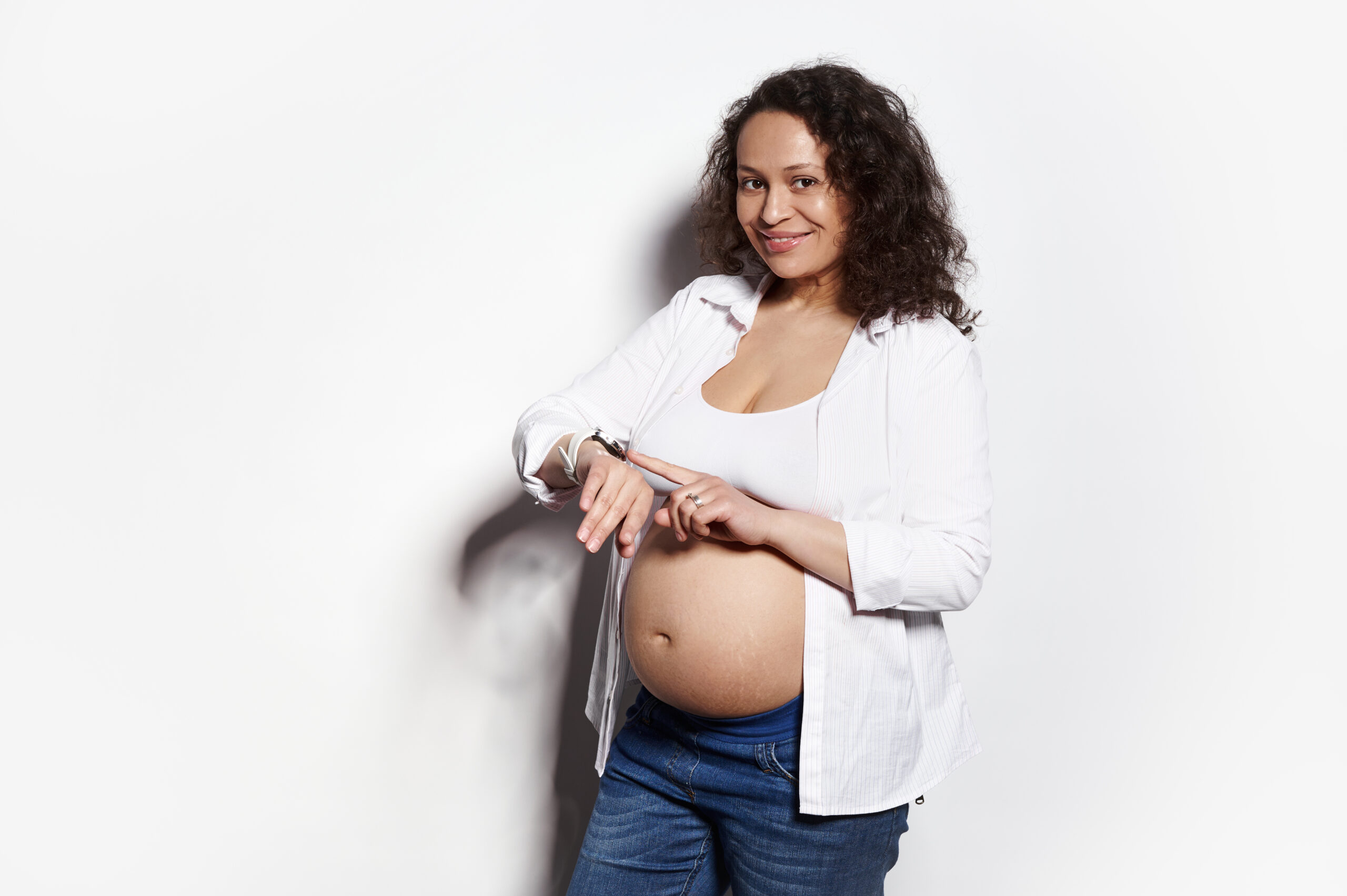 Obrazek ukazuje kobietę w zaawansowanej ciąży, stojącą przed białą ścianą, skupioną na obserwowaniu zegarka na swojej ręce. Jej spojrzenie pełne oczekiwania i niepewności sugeruje, że może to być moment, w którym oczekuje na ważne wydarzenie związane z ciążą, takie jak planowana wizyta u lekarza lub poród.