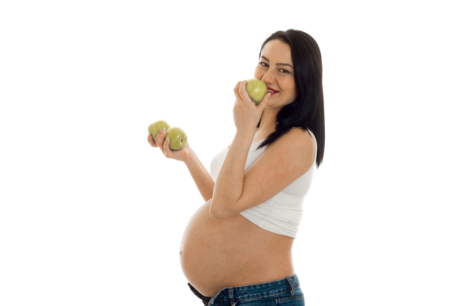 Na tym ujmującym obrazku widzimy kobietę w ciąży, która trzyma w dłoniach kilka zielonych jabłek i zbliża je do ust. Jej uśmiechnięta twarz i promienne spojrzenie sugerują, że owoc ten dostarcza jej wiele przyjemności i korzyści zdrowotnych. Kobieta ubrana jest w wygodną bluzę z długim rękawem, co sugeruje, że jest to zdjęcie zrobione podczas chłodnej pory roku. W tle widać delikatną, pastelową ścianę oraz kilka liści drzewa, co nadaje całej scenie spokojnego i naturalnego charakteru. To piękne zdjęcie idealnie oddaje radość i satysfakcję, jaką kobieta może czerpać z jedzenia zdrowych i smacznych owoców w czasie ciąży.