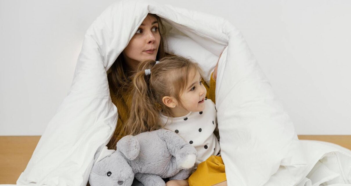 Kobieta z dzieckiem siedzi pod białą bawełnianą kołdrą