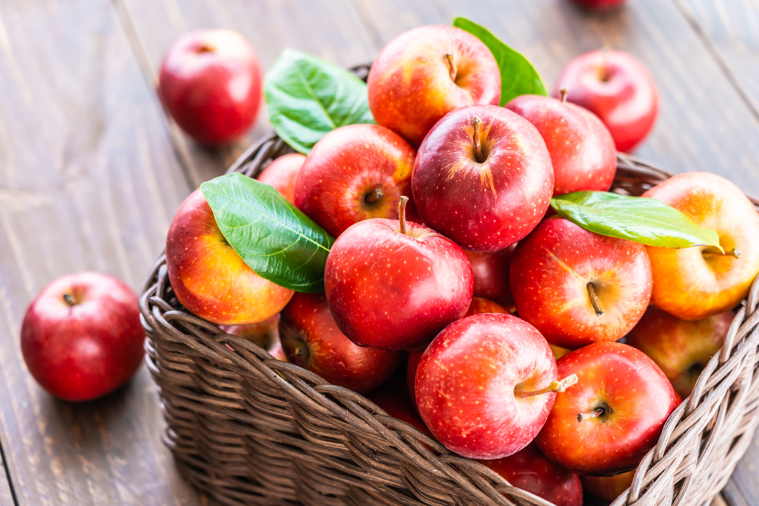 Na obrazku widzimy wiklinowy koszyk, wypełniony czerwonymi jabłkami, ustawiony na blacie. Jabłka wyglądają soczyście i zachęcająco, co może sugerować ich świeżość i smak