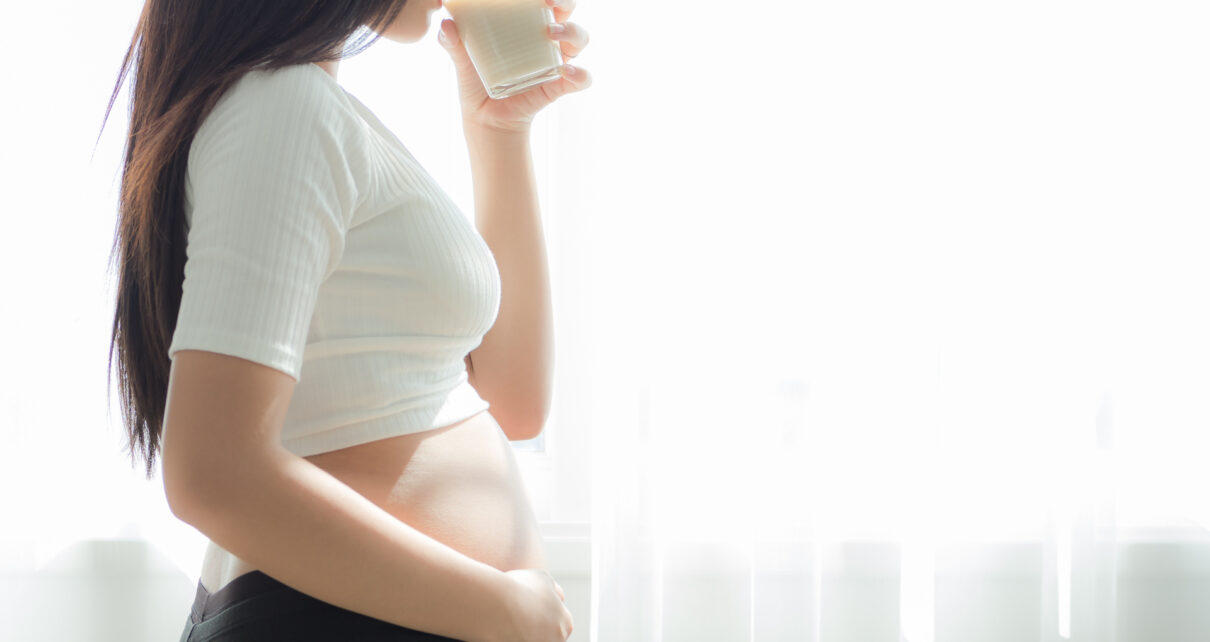 Na zdjęciu widoczna jest kobieta w ciąży, trzymająca w ręku szklankę z napojem. Kobieta patrzy w kierunku szklanki, co sugeruje, że jest skupiona na picu. W tle widać meble oraz ścianę, co wskazuje, że kobieta znajduje się w domu lub w innym prywatnym miejscu. Obrazek ten ma na celu pokazanie, jak ważne jest picie odpowiedniej ilości wody w trakcie ciąży oraz zachęcenie przyszłych mam do dbania o swoje zdrowie i zdrowie dziecka poprzez regularne picie wody i innych napojów.