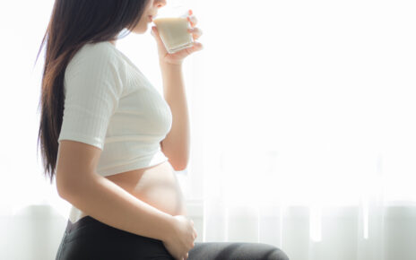 Na zdjęciu widoczna jest kobieta w ciąży, trzymająca w ręku szklankę z napojem. Kobieta patrzy w kierunku szklanki, co sugeruje, że jest skupiona na picu. W tle widać meble oraz ścianę, co wskazuje, że kobieta znajduje się w domu lub w innym prywatnym miejscu. Obrazek ten ma na celu pokazanie, jak ważne jest picie odpowiedniej ilości wody w trakcie ciąży oraz zachęcenie przyszłych mam do dbania o swoje zdrowie i zdrowie dziecka poprzez regularne picie wody i innych napojów.