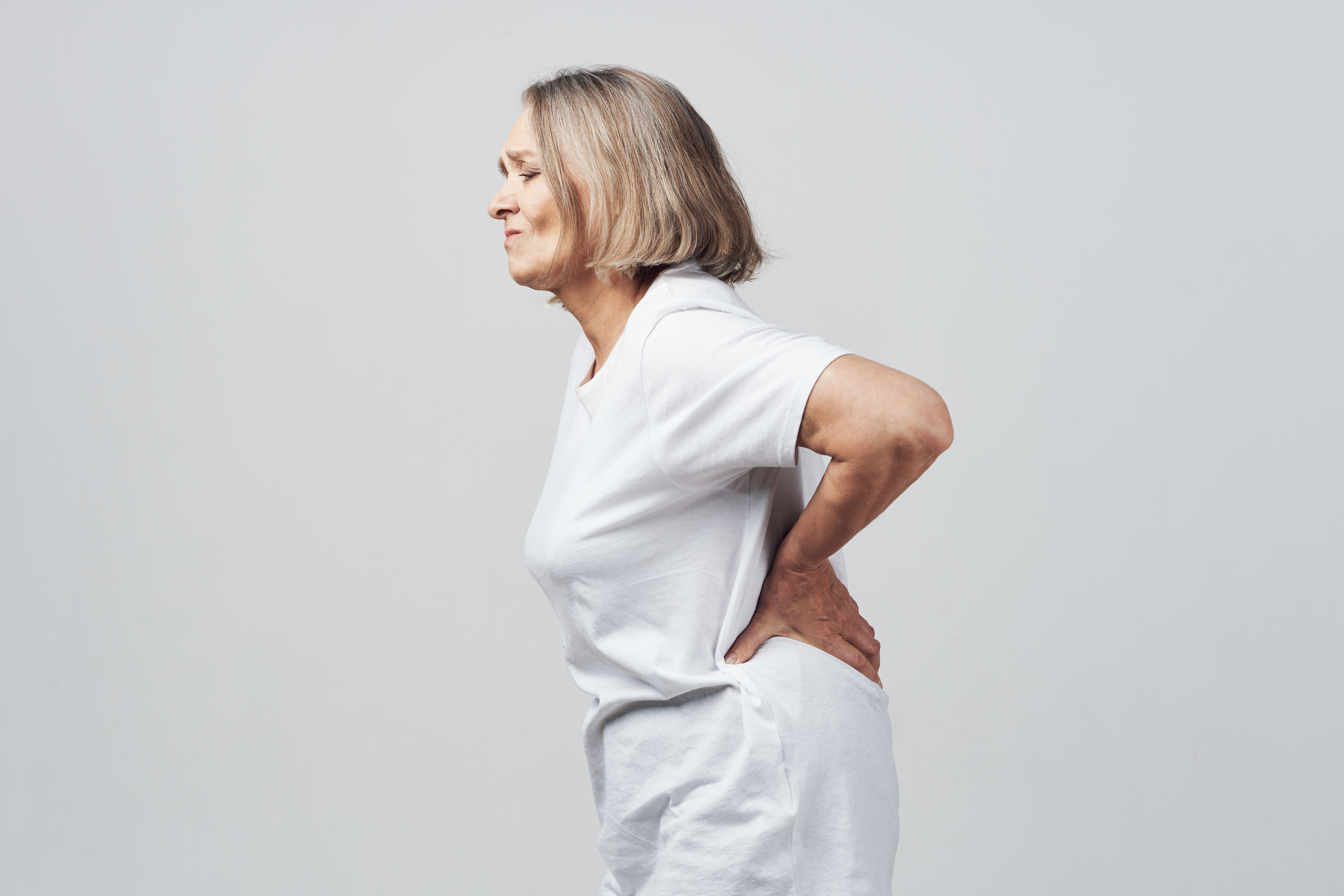 Przedstawione zdjęcie ukazuje starszą kobietę z osteoporozą, która zgina się z bólu pleców. Choroba ta powoduje osłabienie i utratę masy kości, co zwiększa ryzyko złamań i bólu. Kobieta prezentuje się zrezygnowana i cierpiąca, co ukazuje jak trudne i ograniczające mogą być skutki tej choroby